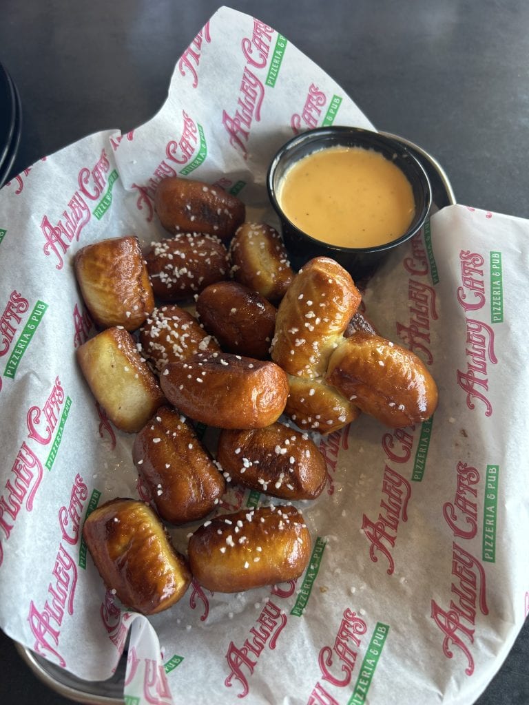 A basket of golden-brown pretzel bites topped with coarse salt, served with a cup of creamy cheese dipping sauce. The basket is lined with branded Alley Cats Pizzeria & Pub paper, and the snack is set on a dark tabletop near the Bagnell Dam Strip.