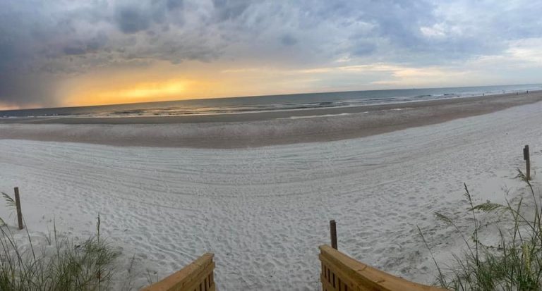 Sunrise on the beach at Amelia Island, with soft golden light breaking through dark clouds over the calm Atlantic Ocean. Wooden steps lead down to wide, white sand bordered by gentle waves, capturing a peaceful coastal moment.