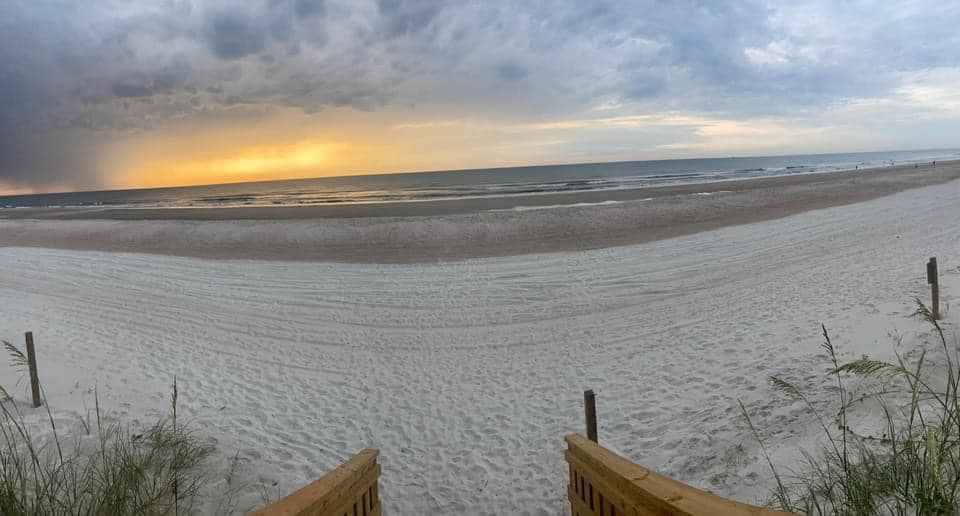 Panoramic view of a wide, empty beach on Amelia Island at dusk, with soft golden light breaking through a cloudy sky over the ocean. Wooden dune stairs lead down to smooth, white sand, with sea oats framing the foreground.