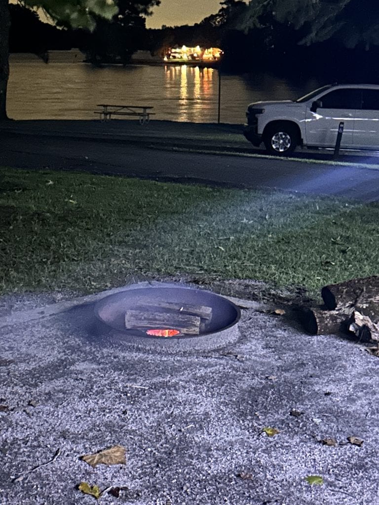 A campfire ring with glowing embers sits in the foreground of a nighttime lakeside scene. Across the water, a brightly lit boat reflects on the surface, while a white pickup truck is parked nearby and a picnic table rests along the shoreline.
