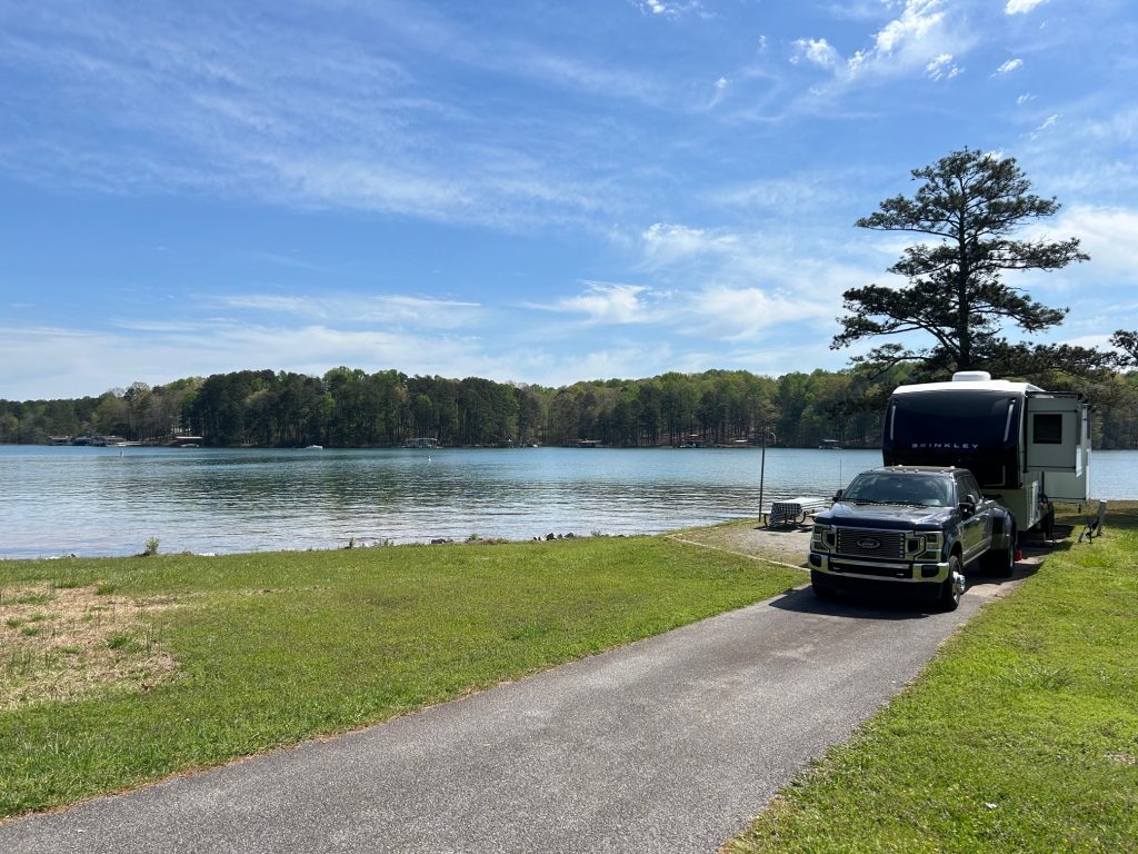 Lakeside RV campsite at Bald Ridge Campground with a large Brinkley RV and black Ford truck parked on a paved drive beside calm water. The site includes a picnic table and grill on a concrete pad, surrounded by green grass and views of a forested shoreline under a partly cloudy sky.