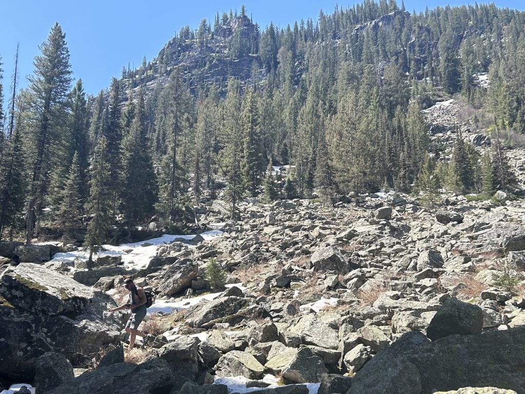 Alan navigates a rugged, rocky section of the Jenny Lake hike to Inspiration Point, with patches of snow still visible among the boulders. Towering pine trees and a steep, forested mountainside rise in the background under a clear blue sky.