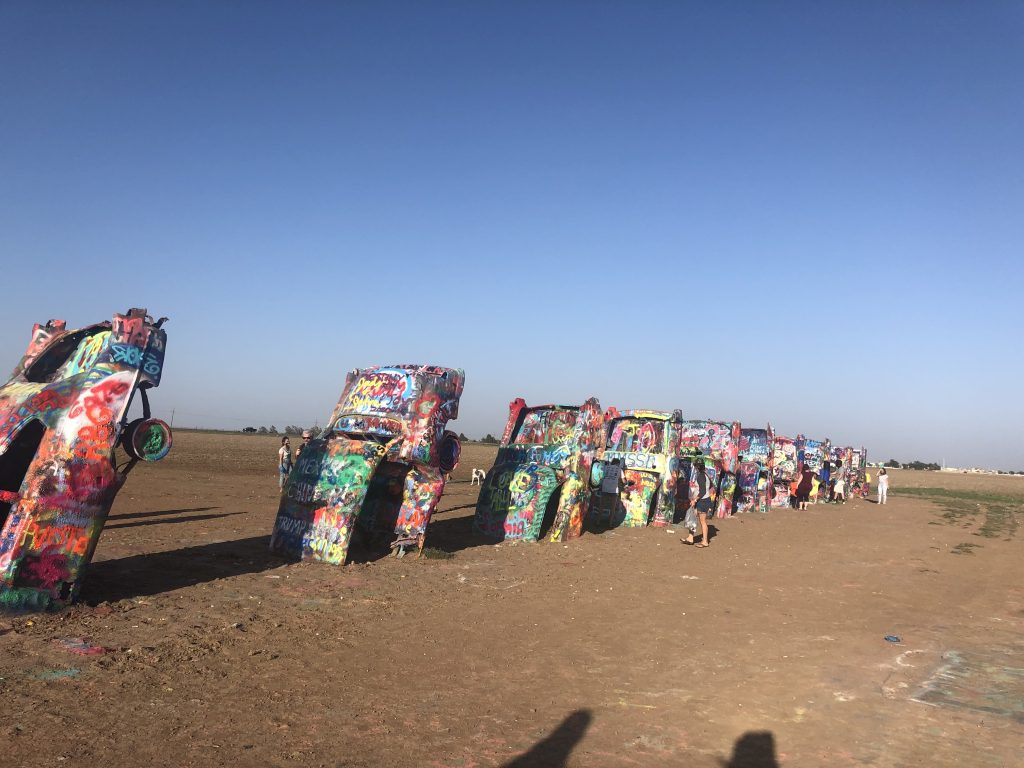 Half-buried vintage cars covered in layers of colorful spray paint stand in a row in a flat, dusty field. Visitors walk among the vibrant art installation, leaving fresh graffiti under a clear blue sky.