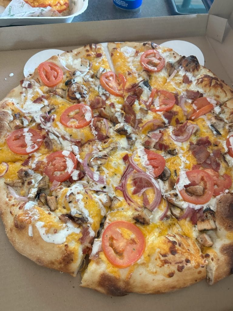 Close-up of a freshly baked pizza topped with sliced tomatoes, red onions, grilled chicken, bacon, cheddar cheese, and a drizzle of ranch dressing. The pizza sits in an open box, with a cheesy sandwich visible in the background.