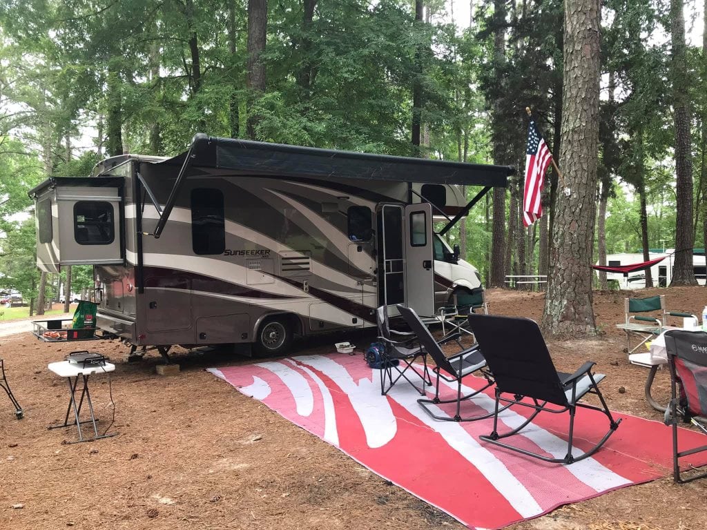 A Sunseeker Class C motorhome is parked at a wooded campsite with its awning extended over a large red and white outdoor mat. Folding chairs, tables, and an American flag hanging from a nearby tree create a relaxed camping setup among tall pine trees.
