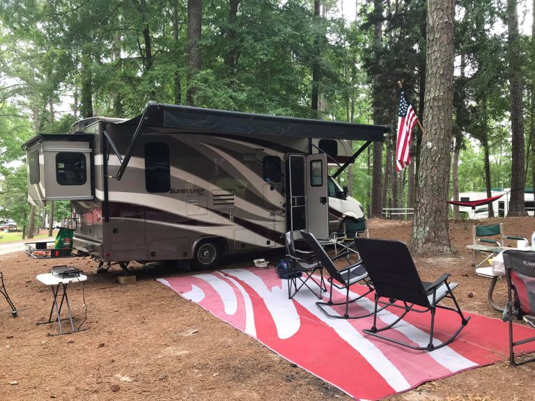 A Sunseeker Class C motorhome is parked at a wooded campsite with its awning extended over a large red and white outdoor mat. Folding chairs, tables, and an American flag hanging from a nearby tree create a relaxed camping setup among tall pine trees.