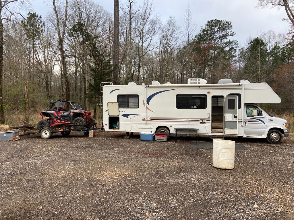 A Class C motorhome is parked at a gravel campsite with its door open and a red side-by-side off-road vehicle loaded on a trailer behind it, surrounded by bare trees on an overcast day.