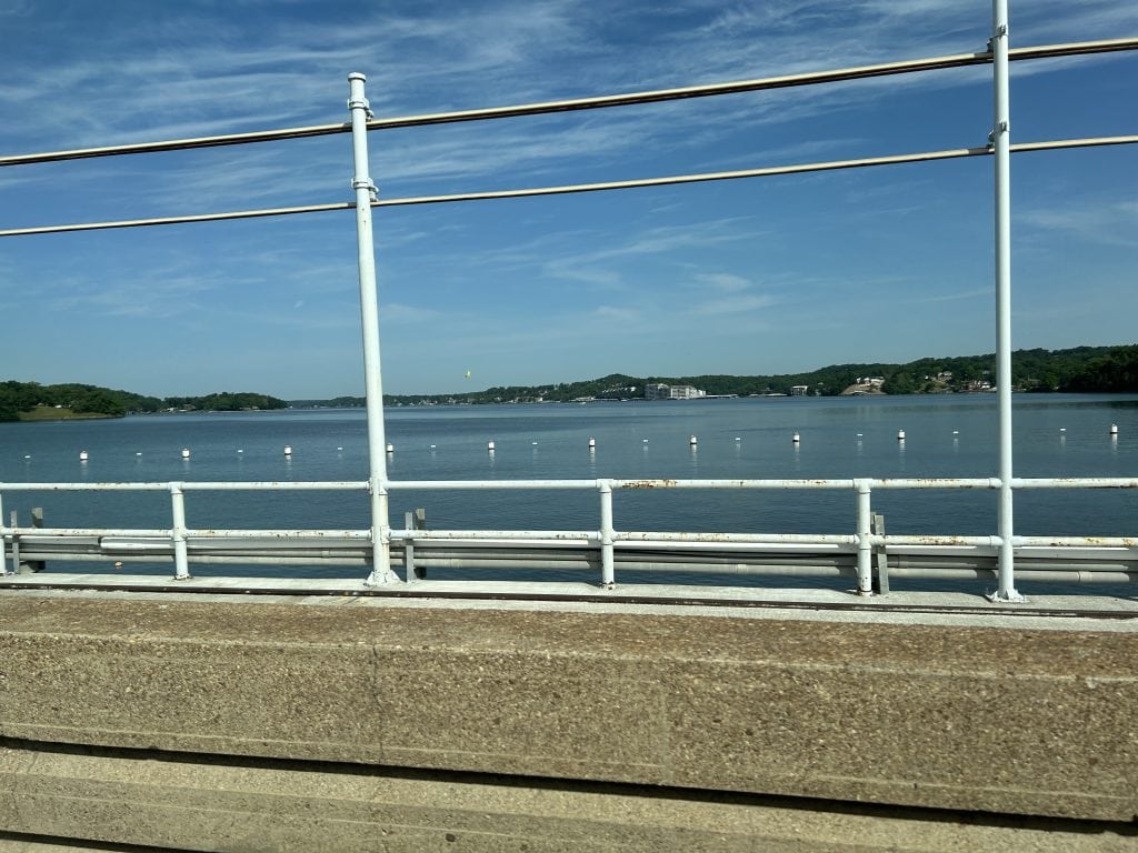 View across Bagnell Dam, with guardrails in the foreground and the calm waters of Lake of the Ozarks stretching into the distance. Forested hills and buildings line the far shore under a bright blue sky with wispy clouds.