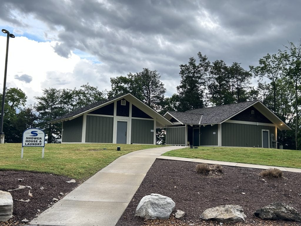 Two modern green and white buildings labeled as the shower house and laundry facility at Elm Hill RV Resort. A paved walkway leads uphill to the buildings, with a small lawn sign marking the area, surrounded by trees and under a cloudy sky.