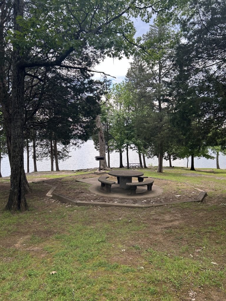 Shaded lakeside picnic area at Elm Hill RV Resort with a round concrete table and curved benches surrounded by trees. A metal grill and additional picnic table are set closer to the water, with views of the lake visible through the tree line.