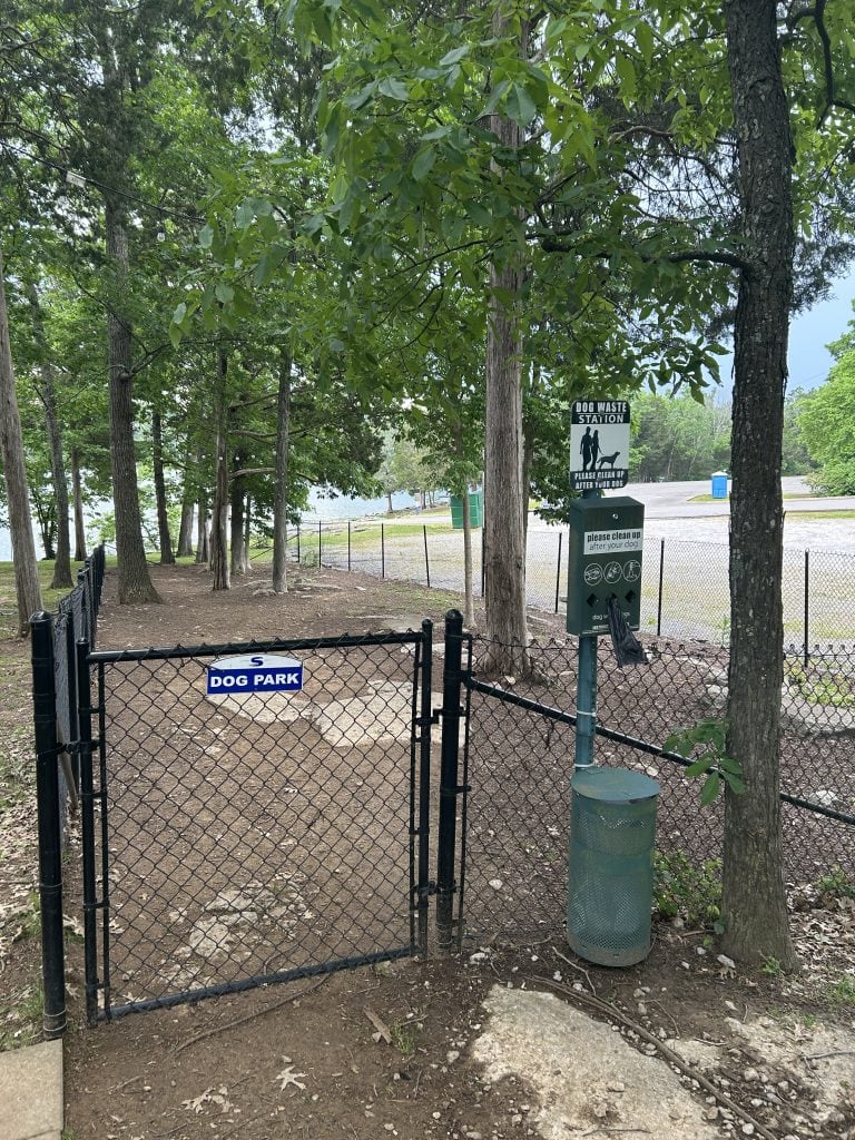Gated entrance to the dog park at Elm Hill RV Resort, surrounded by tall trees with a dog waste station and trash can beside the fence. The area is enclosed with chain-link fencing and has a dirt surface with scattered leaves.