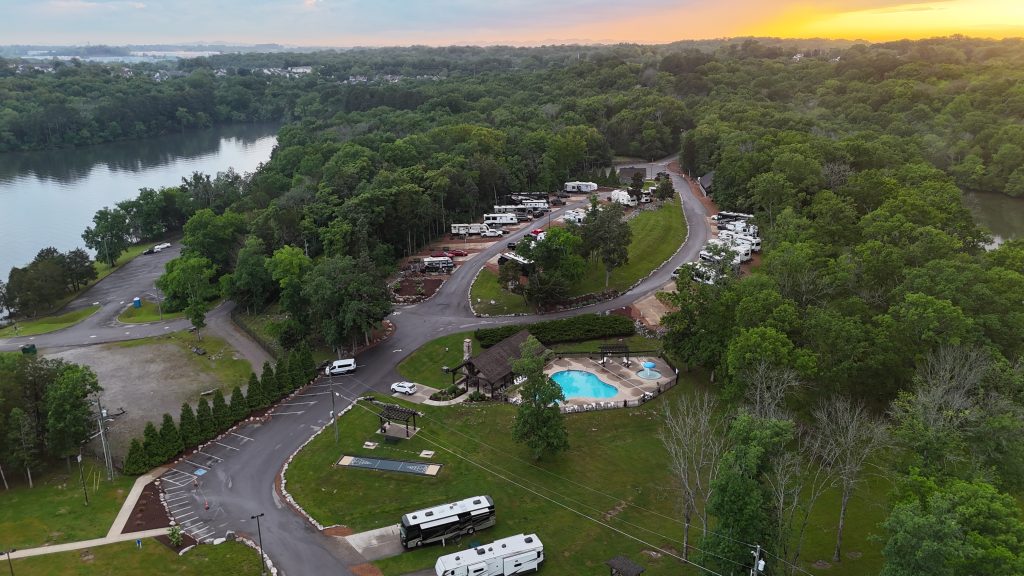 Aerial view of Elm Hill RV Resort nestled among dense green trees beside a peaceful lake at sunset. The resort features rows of RVs parked along winding roads, a fenced swimming pool area with a hot tub, a shuffleboard court, and a community pavilion. The lake and surrounding forest create a serene natural setting, with golden light from the setting sun illuminating the horizon.