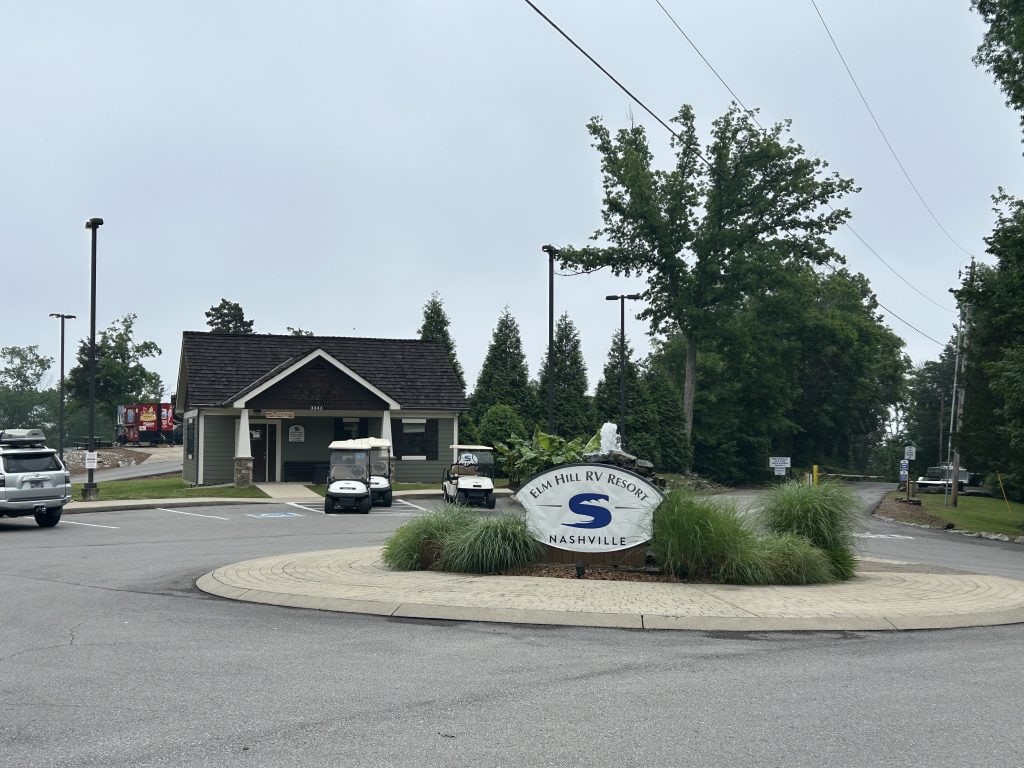 Front entrance to Elm Hill RV Resort in Nashville featuring a roundabout with a landscaped sign displaying the resort’s name and logo before you arrive at the gates. A small office building with a wooden roof sits nearby, with golf carts parked out front and a gated road leading into the tree-lined campground.