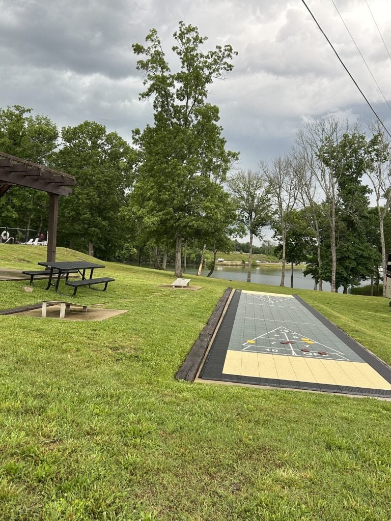 Outdoor shuffleboard court set on a grassy lawn near a lake, with game pieces arranged on the playing surface. A black metal picnic table and wooden bench sit nearby under cloudy skies, surrounded by trees and peaceful scenery.