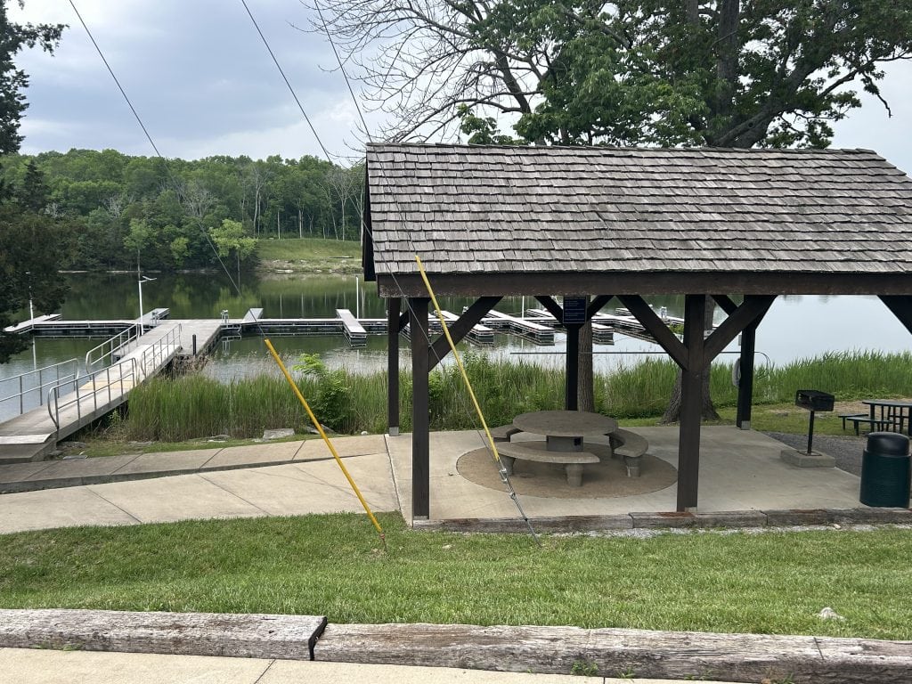 Covered picnic pavilion with a round concrete table and benches overlooking a calm lake with boat docks and ramps. The area is surrounded by green grass, trees, and reeds, creating a peaceful waterfront setting.