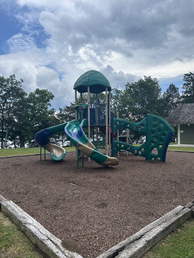 Colorful playground structure with multiple slides, climbing walls, and a green dome roof set on a mulch surface. Surrounded by grassy areas and trees, the playground overlooks a wooded lakeside under a partly cloudy sky.
