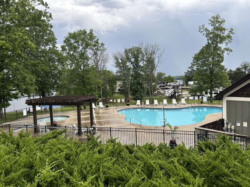 Fenced-in outdoor pool area at Elm Hill RV Resort featuring a large kidney-shaped swimming pool, a smaller hot tub under a wooden pergola, and rows of white lounge chairs. Surrounded by green trees and RVs, the space overlooks a lake under a cloudy sky.