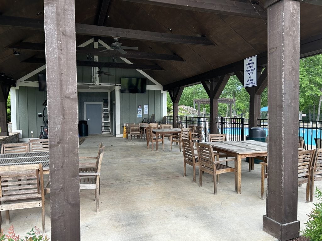 Covered outdoor patio with wooden tables and chairs next to a fenced pool area at Elm Hill RV Resort. Ceiling fans, mounted TVs, and a sign reading “No drinks, food, coolers, pets or glass allowed” enhance the shaded lounging space.