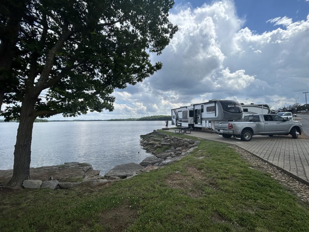 A silver pickup truck and an Alliance Valor fifth-wheel RV are parked beside a calm lakeshore under a partly cloudy sky. A large tree shades the rocky shoreline in the foreground, with a picnic table and a person standing near the water’s edge.