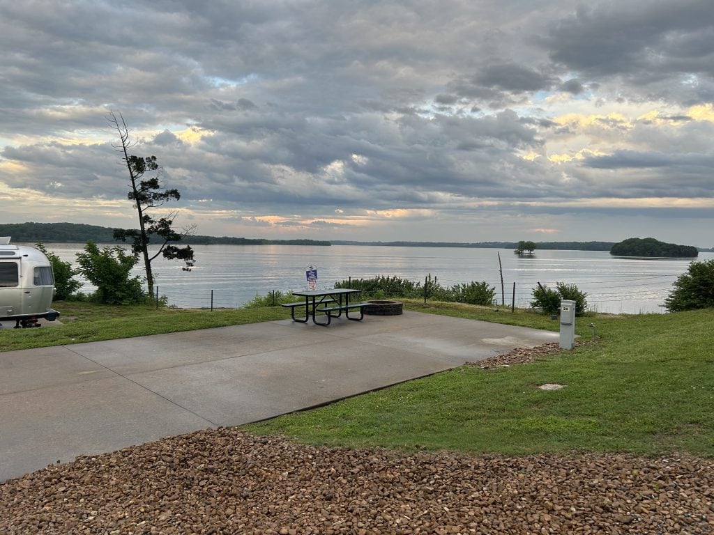 Lakeside RV campsite with a concrete pad, picnic table, and fire ring overlooking calm water and a distant tree-covered island. Airstream trailer is partially visible on the left, with dramatic clouds and soft evening light in the sky.