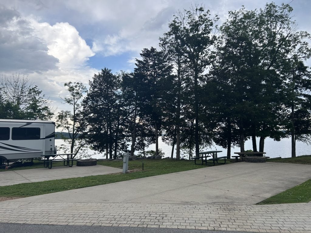 Standard lakeside RV campsites at Elm Hill RV Resort with concrete pads, picnic tables, and fire rings at a wooded campground near J. Percy Priest Lake. Trees partially obscure the water, and a large RV is parked to the left under a partly cloudy sky.