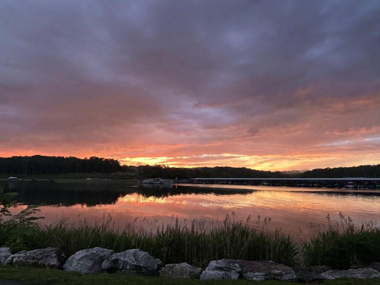 Vibrant sunset over J. Percy Priest Lake, with fiery orange and yellow hues reflecting on the calm water. Silhouetted hills and a marina appear in the distance, while rocks and tall grass line the lake’s edge in the foreground, creating a peaceful and dramatic scene.