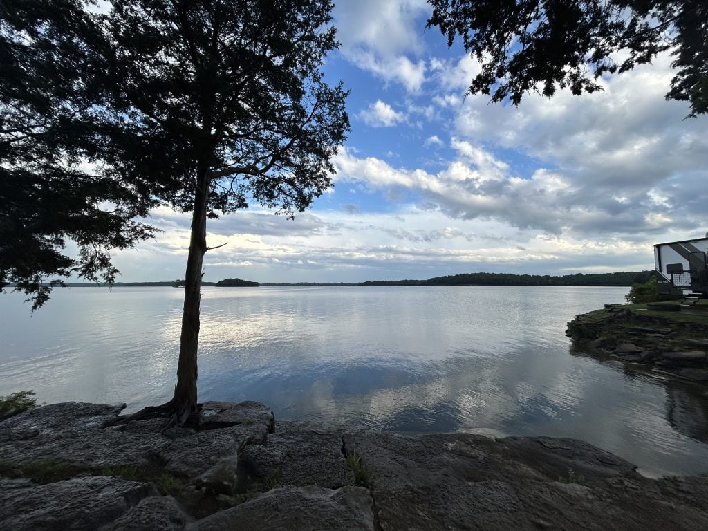Peaceful lakeside view at Elm Hill RV Resort with calm water reflecting a partly cloudy sky. A lone tree stands on rocky shoreline in the foreground, with a modern RV parked near the edge and a small tree-covered island visible in the distance.
