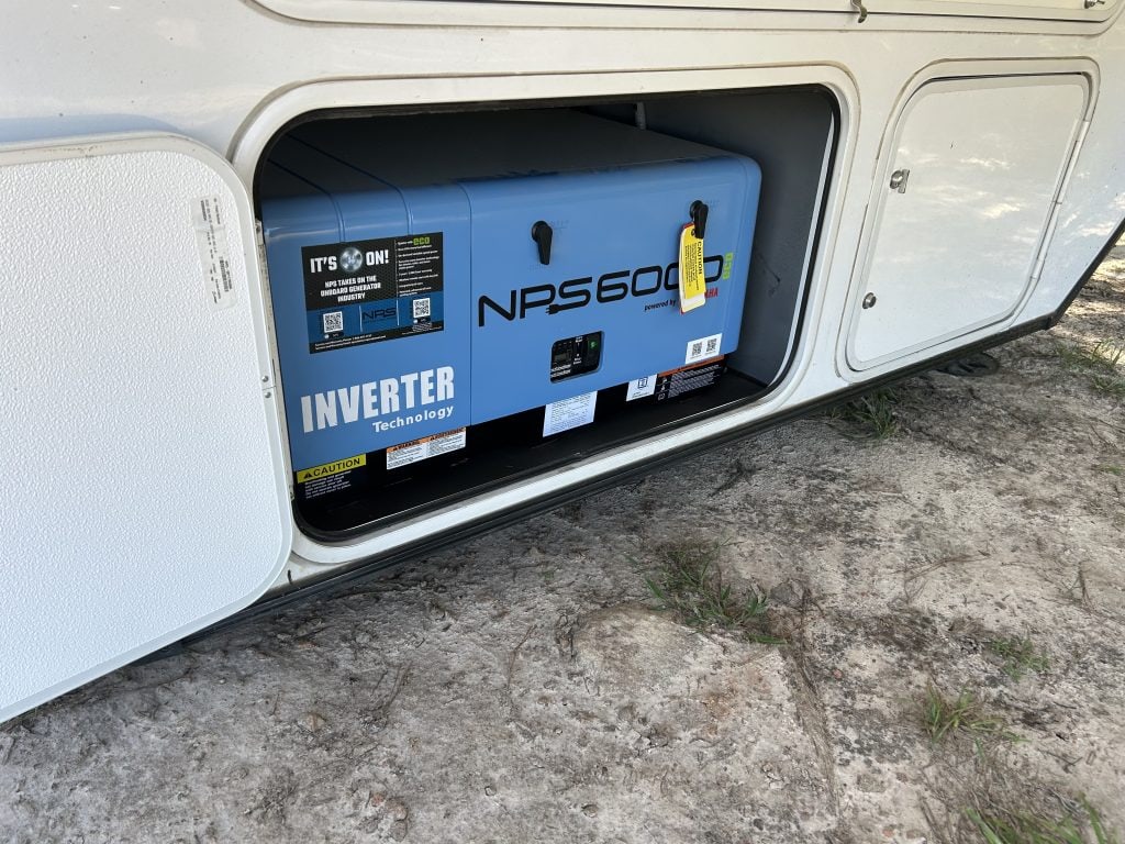 A blue NPS6000 inverter generator is stored inside a side compartment of an RV. The compartment door is open, revealing caution labels and a yellow tag attached to the generator handle.
