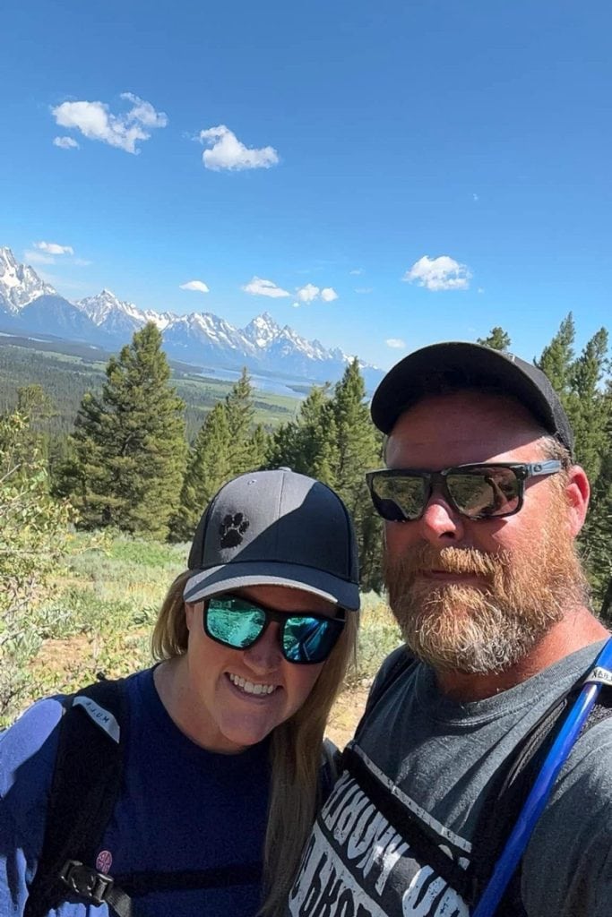 Morgan and Alan pose for a selfie while hiking in Grand Teton National Park, with pine trees and snow-capped mountains in the background under a clear blue sky.