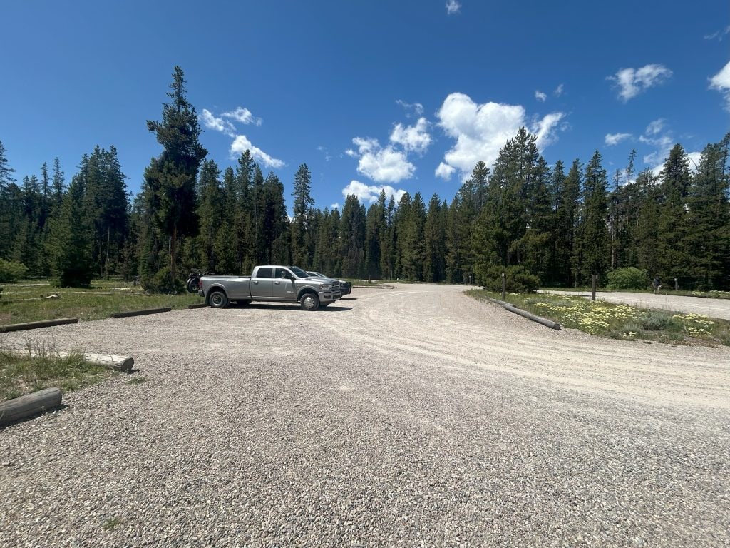 A nearly empty gravel parking lot sits under a bright blue sky, with a single silver pickup truck parked near the center. Tall evergreen trees line the lot’s edge, creating a forested backdrop.