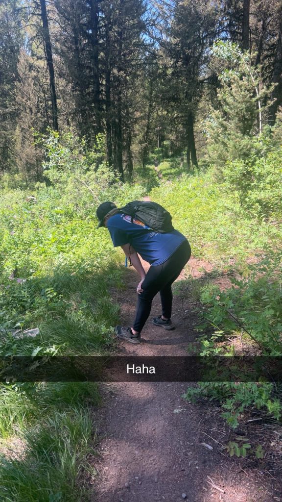 Morgan, a woman, wearing a backpack, navy shirt, and black leggings bends forward to rest on a shaded forest trail surrounded by dense green foliage. Sunlight filters through tall trees, creating patches of light along the dirt path.