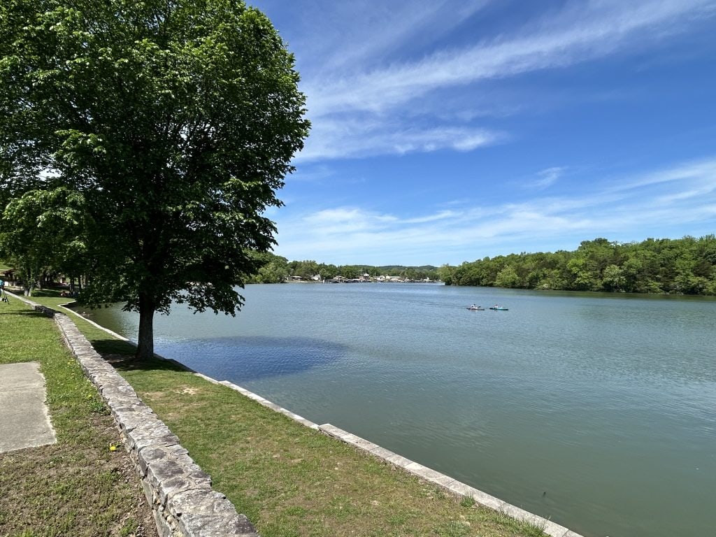 A peaceful lakeside scene at Ha Ha Tonka State Park in Missouri, with a stone wall lining the grassy shoreline. A large tree offers shade on the left, while a calm, blue lake stretches out under a partly cloudy sky. A few people are kayaking or paddleboarding in the distance, surrounded by lush green forest.