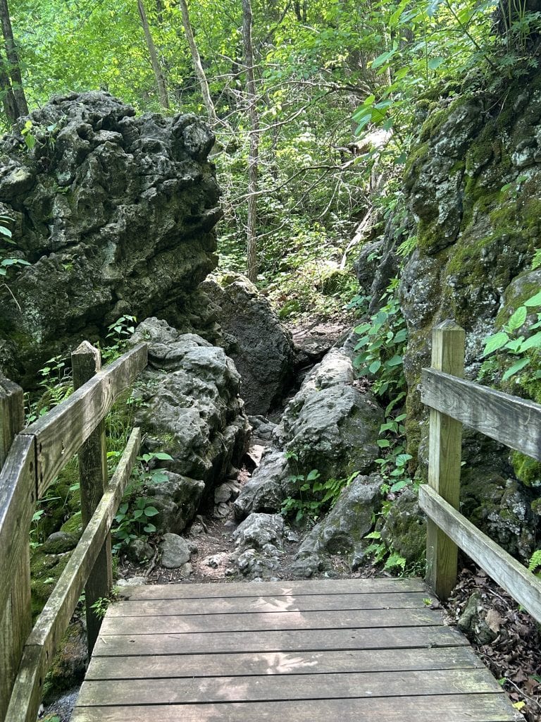 A wooden footbridge leads into a rugged, rocky section of the Natural Spring Trail at Ha Ha Tonka State Park in Missouri. Large moss-covered boulders frame a narrow path ahead, surrounded by dense green foliage under the forest canopy.