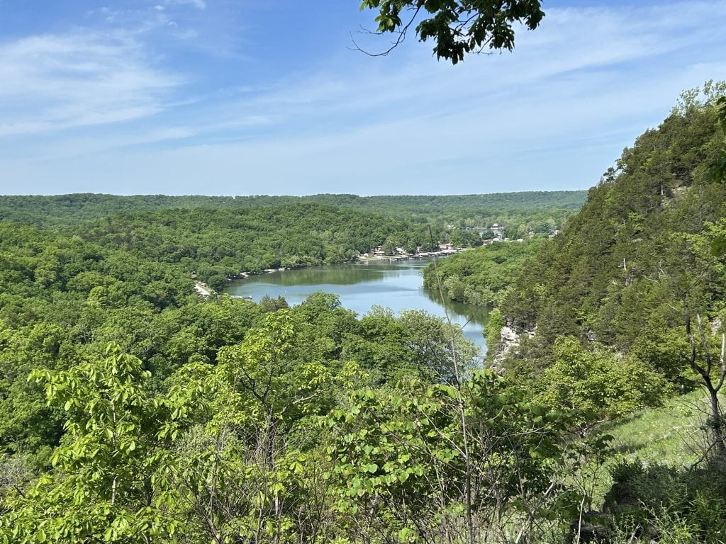 Scenic overlook at Ha Ha Tonka State Park showing a lush green valley and a winding lake surrounded by dense forest. The view is taken from near the castle ruins, with clear blue skies and distant buildings nestled along the shoreline.