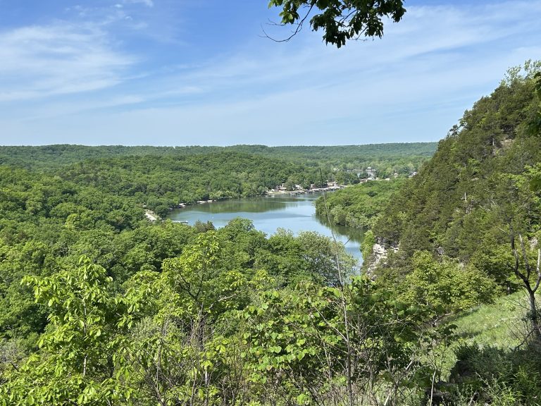Scenic overlook at Ha Ha Tonka State Park showing a lush green valley and a winding lake surrounded by dense forest. The view is taken from near the castle ruins, with clear blue skies and distant buildings nestled along the shoreline.