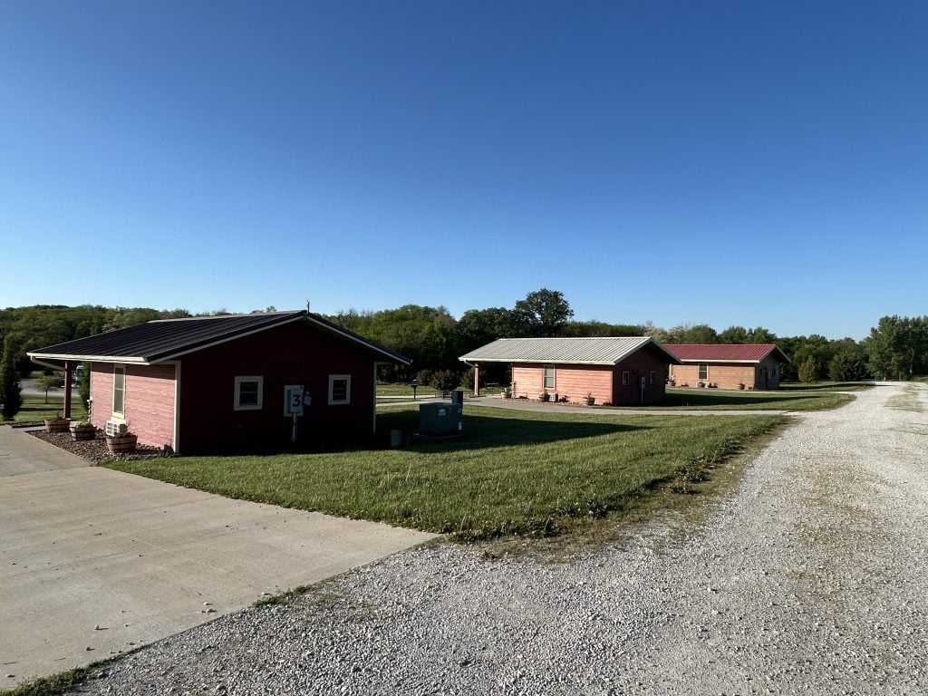 Row of small red cabins at Hidden Falls Cabins & RV Park with cabin 3 closest to the camera, each set on a well-maintained lawn beside a gravel road. The scene is quiet under a clear blue sky, with trees lining the background.