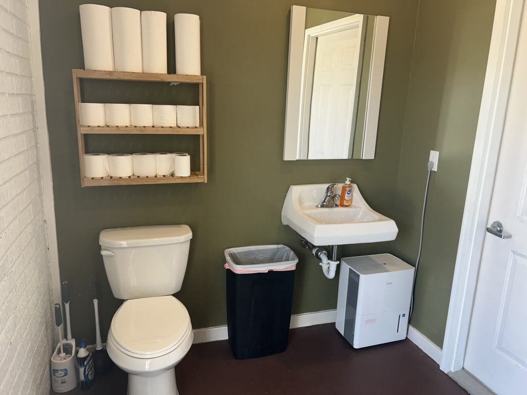 Clean restroom at Hidden Falls Cabins & RV Park featuring a toilet, a wall-mounted sink with liquid soap, and a mirror above it. A wooden shelf holds extra toilet paper and paper towels, with a trash can and dehumidifier placed below.