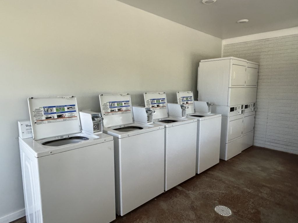 Laundry room at Hidden Falls Cabins & RV Park featuring four top-loading washing machines and two stacked washer-dryer units against a white and brick wall. The space has a clean, open layout with a concrete floor and floor drain.