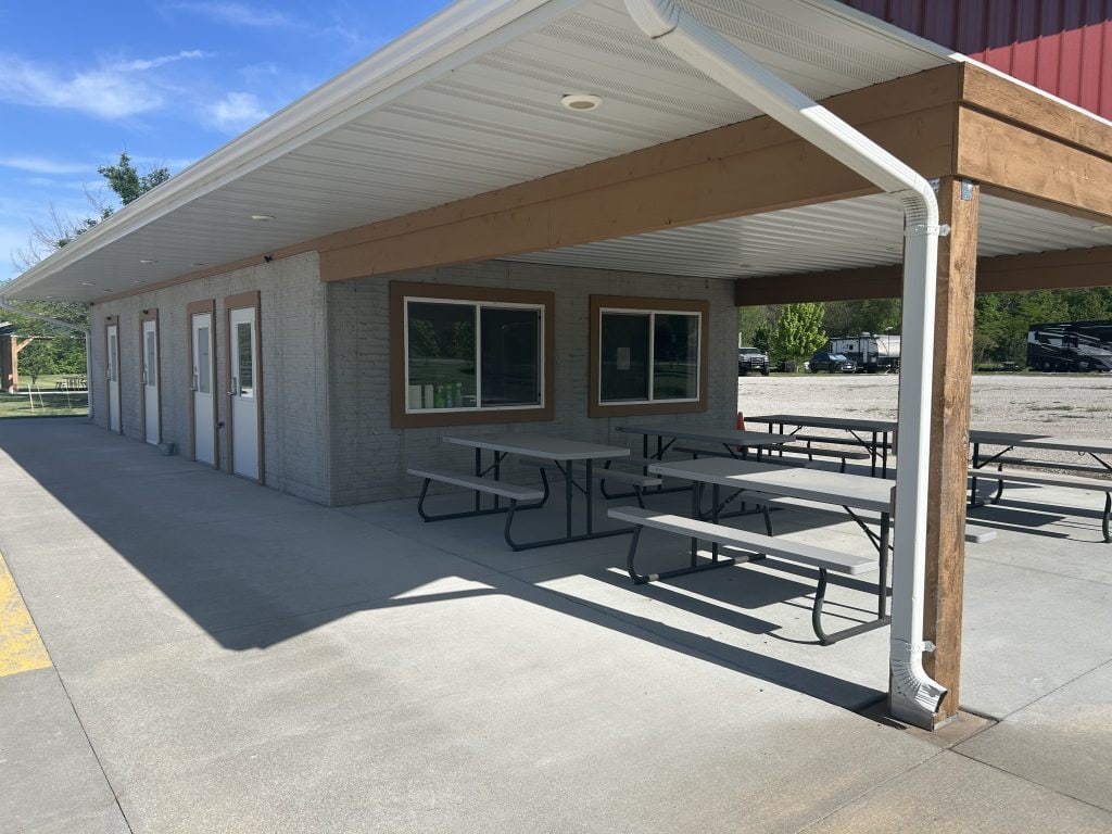 Covered outdoor seating area at Hidden Falls Cabins & RV Park with several picnic tables beneath a white metal roof supported by wooden posts. The building features multiple doors and a large window, with RVs visible in the gravel lot beyond.
