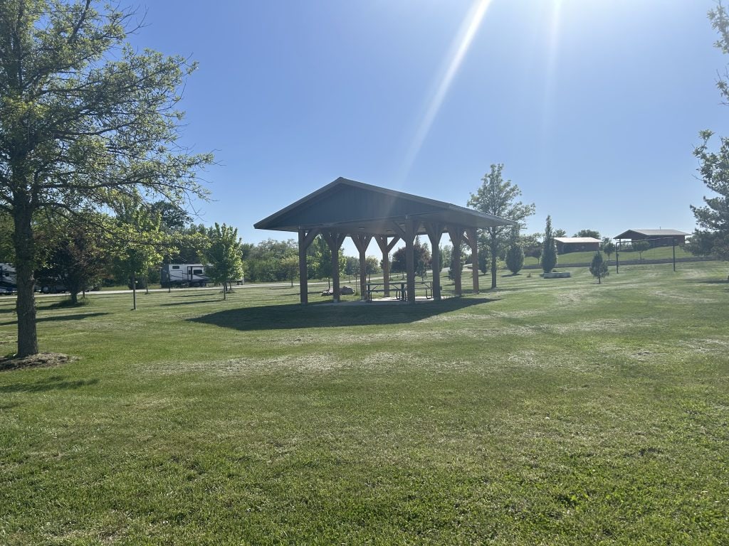 Grassy field at Hidden Falls Cabins & RV Park featuring a wooden picnic shelter with a metal roof, surrounded by trees, open space, and distant RVs and cabins. Sunlight beams through a clear blue sky, casting long shadows across the lawn.