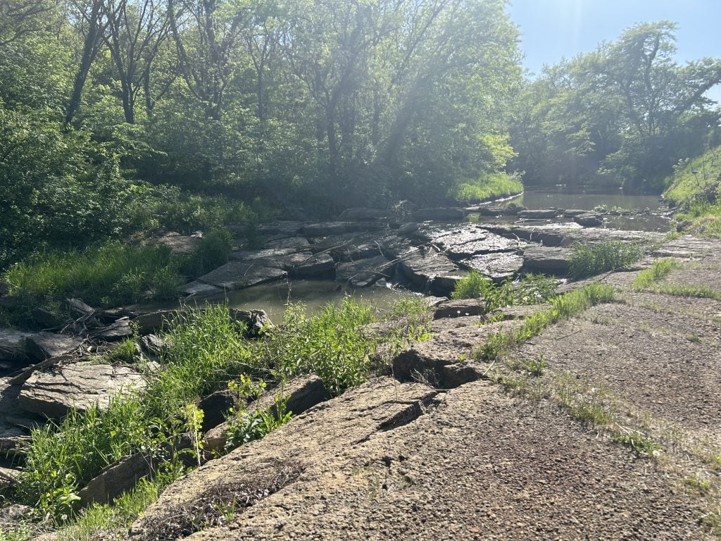 Shaded woodland creek at Hidden Falls Cabins & RV Park, with layered flat rocks forming a natural cascade and patches of green grass and plants growing between them. Sunlight filters through the trees and reflects off the water’s surface.