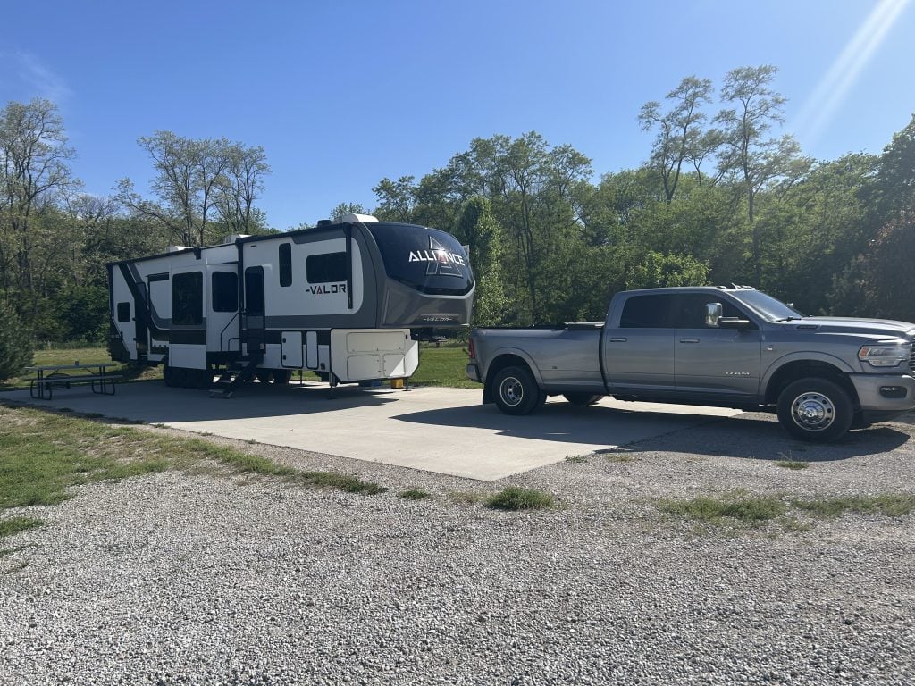 A silver dually pickup truck is parked in front of a white, black, and gray Alliance Valor fifth-wheel RV parked on a concrete pad in a sunny campground. The site is surrounded by green trees, with a picnic table visible beside the RV.