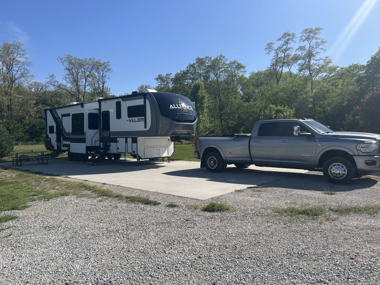 A silver dually pickup truck is parked in front of a white, black, and gray Alliance Valor fifth-wheel RV parked on a concrete pad in a sunny campground. The site is surrounded by green trees, with a picnic table visible beside the RV.