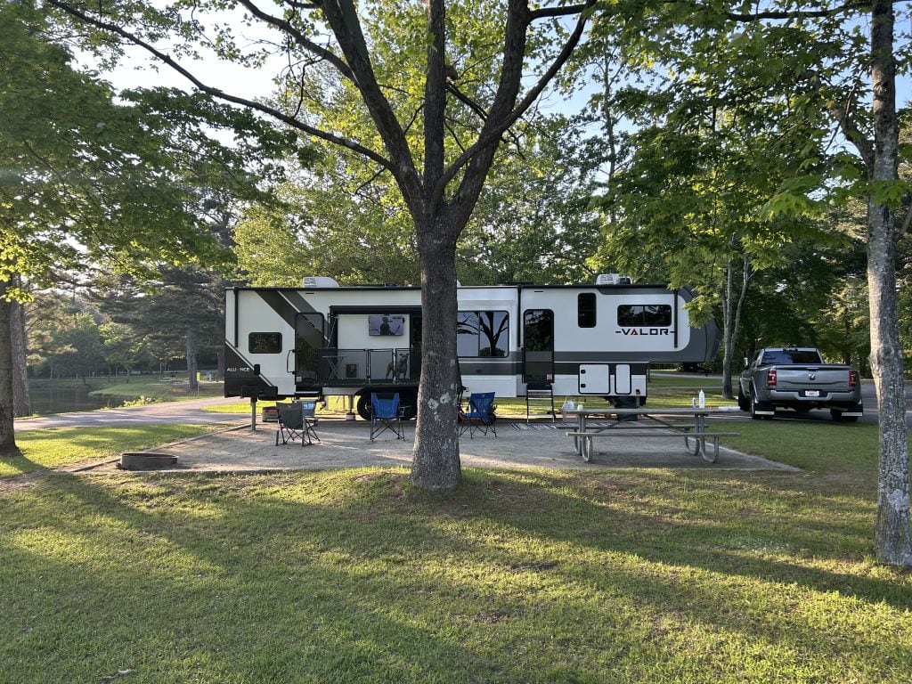 Spacious RV campsite at Bald Ridge Campground with a large Valor fifth-wheel trailer, camping chairs, picnic table, and fire ring set on a gravel pad beneath shady trees. A TV is mounted outside the RV and a gray pickup truck is parked nearby on the paved drive.