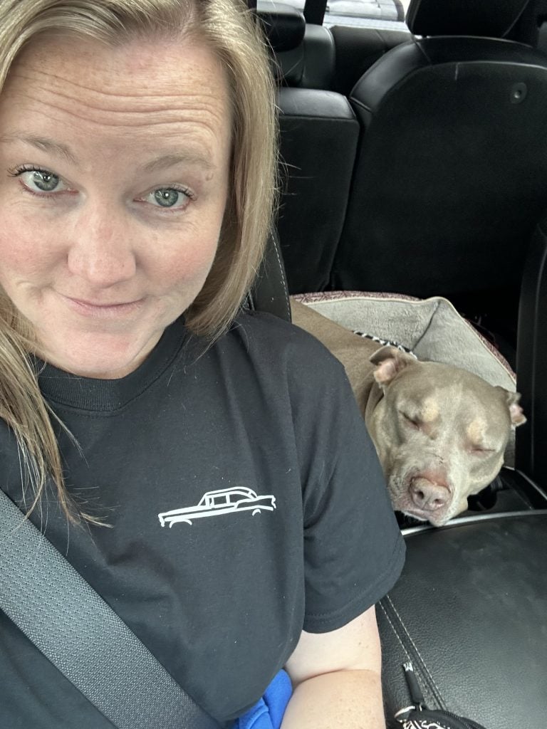 Morgan takes a selfie in the front seat of a vehicle while Coco, a large tan dog, sleeps peacefully in a bed on the backseat, capturing a relaxed travel moment.