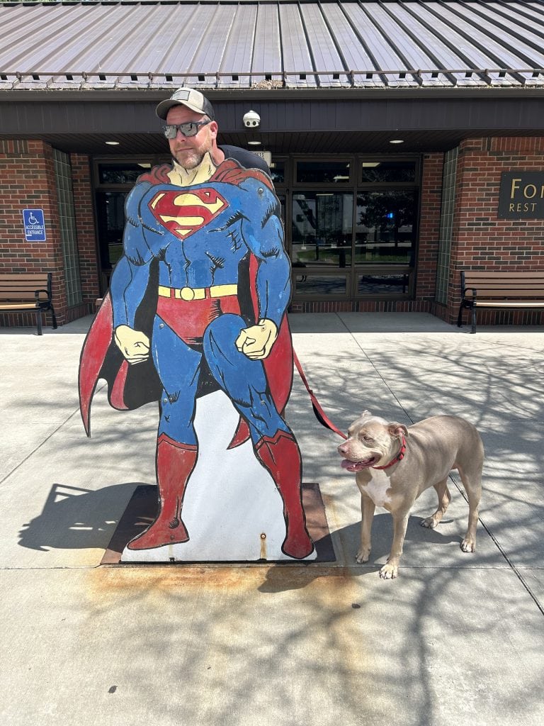 Alan poses with his face through a Superman cutout in front of a rest area building, holding the leash of a smiling tan dog standing beside him on the sunny sidewalk.