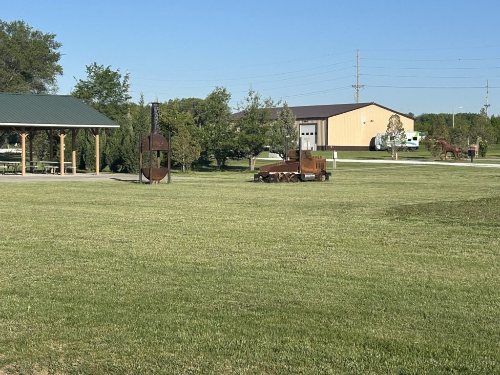 Open grassy area at Hidden Falls Cabins & RV Park featuring a wooden picnic shelter, a tall metal chiminea, and rustic metal sculptures including a tractor and a horse. A tan metal building and RV are visible in the background under a clear blue sky.