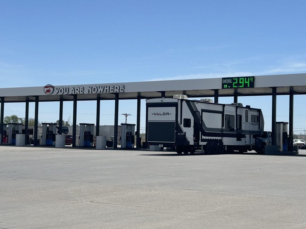 A large fifth wheel RV is parked at a gas station pump under a canopy with a sign reading “YOU ARE NOWHERE.” An electronic display shows diesel priced at $2.94 per gallon against a clear blue sky.