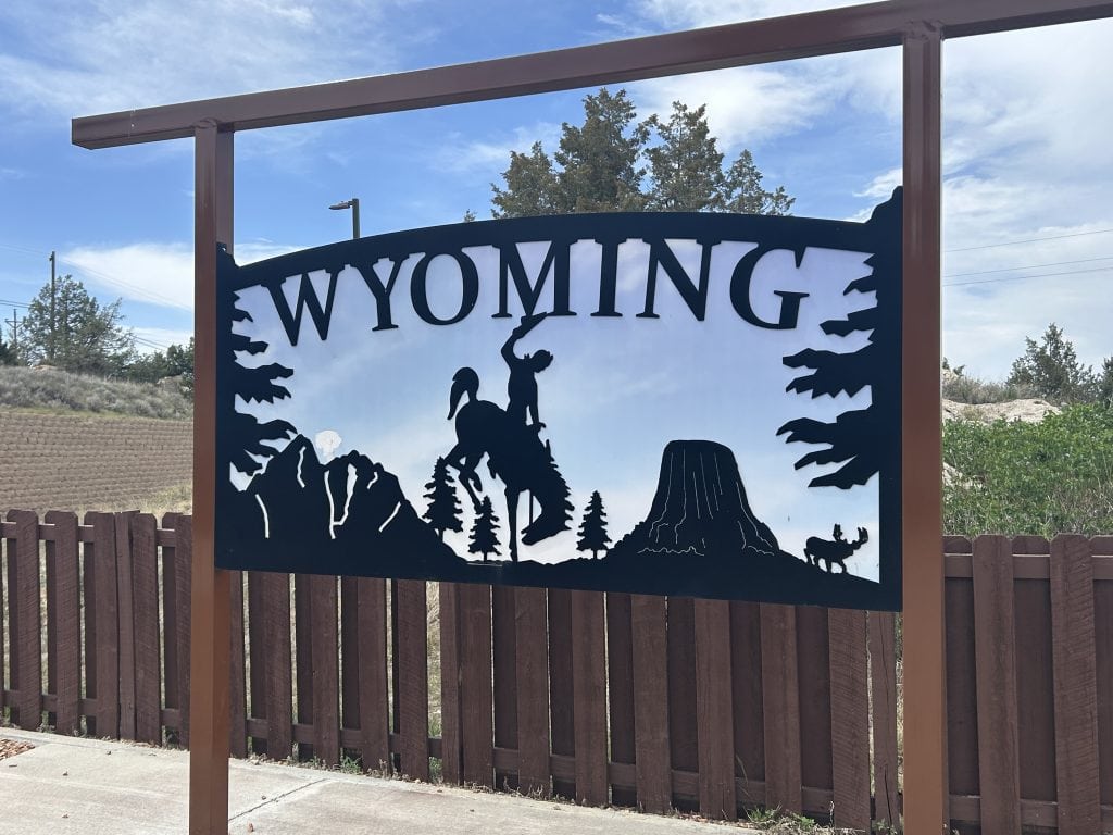 A decorative metal Wyoming sign features silhouettes of a bucking horse and rider, mountain peaks, pine trees, Devil’s Tower, and a moose, set against a blue sky at a roadside rest stop in Wyoming.