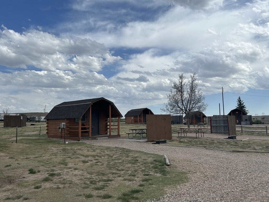 Row of rustic wooden cabins at Cheyenne RV Resort by RJourney, each with a porch, picnic table, and nearby privacy fences. The cabins sit on gravel paths surrounded by sparse grass under a partly cloudy sky.